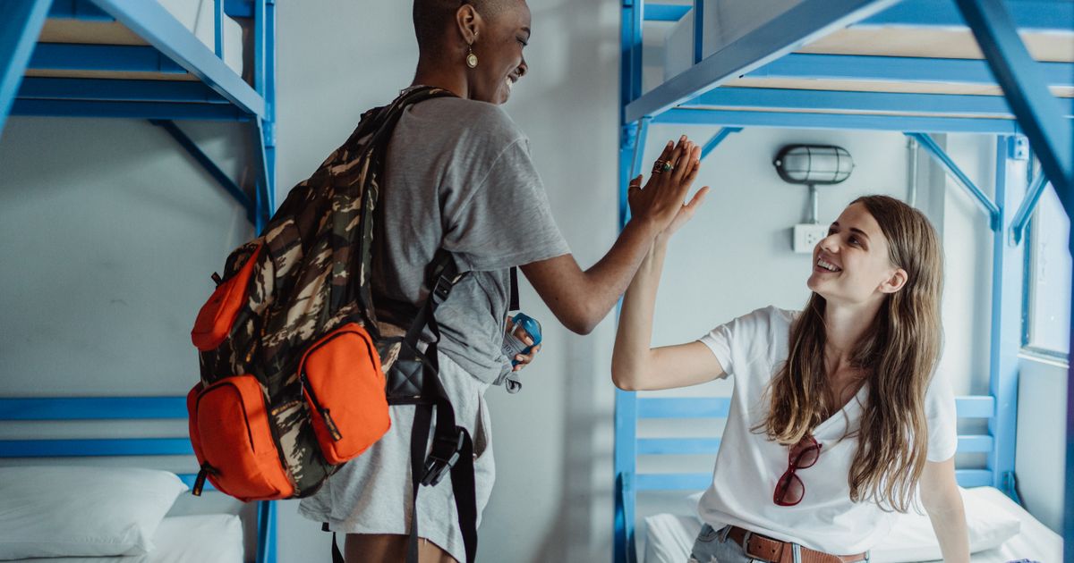 Two female backpackers high-fiving in a bright hostel dorm room, enjoying travel adventures.