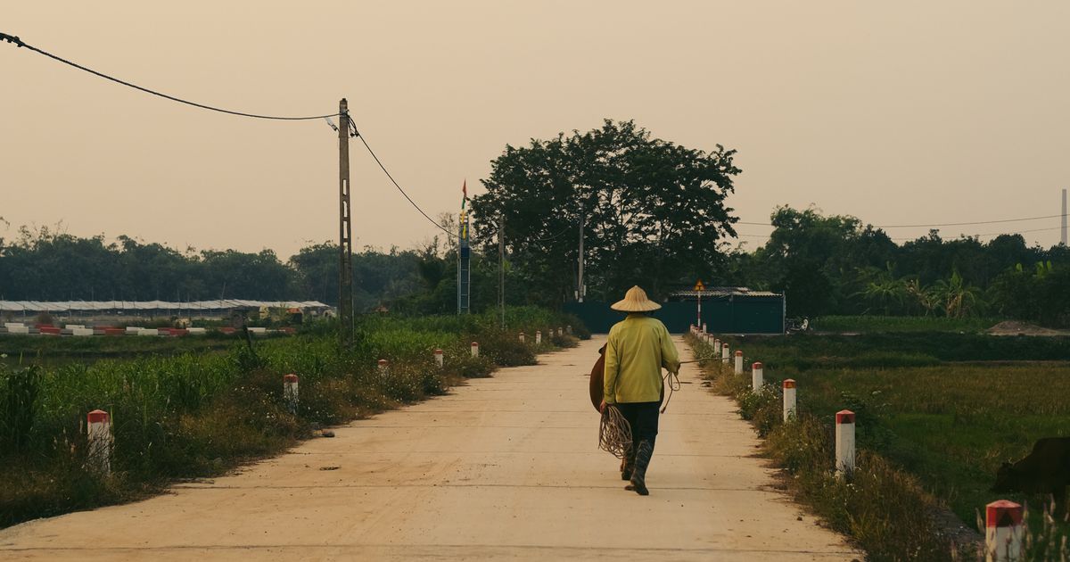 A person in traditional attire walks along a rural path in Hà Nam, Vietnam, with lush greenery.